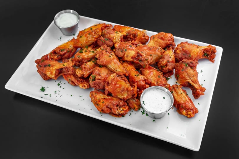 A large platter of buffalo chicken wings with two small cups of ranch dressing.
