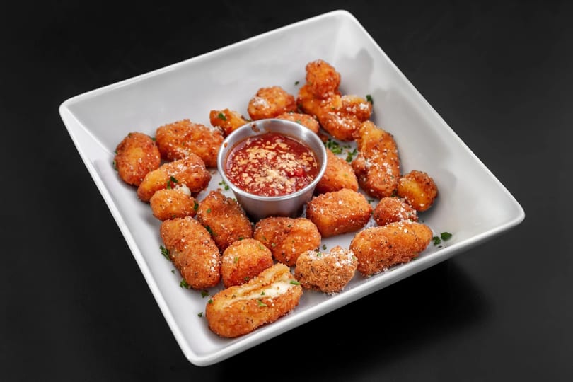 A white rectangular plate filled with golden-brown fried mozzarella bites and a side of marinara sauce.