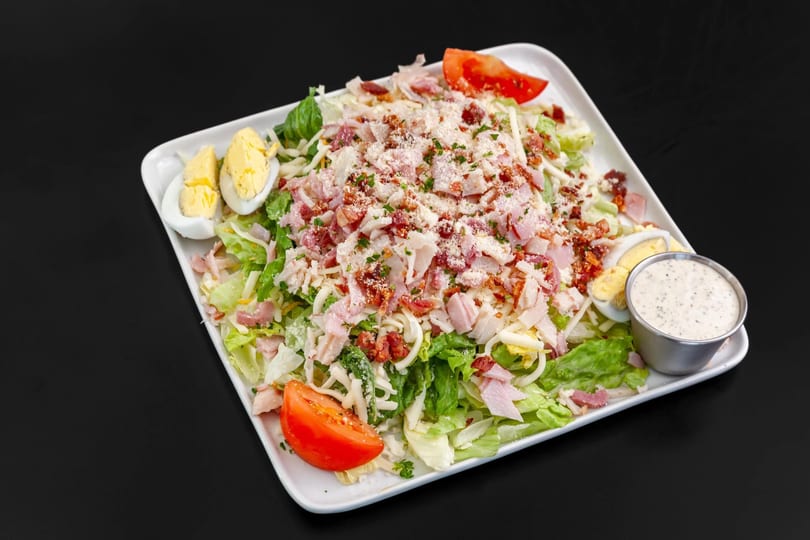 A large chef salad with lettuce, ham, bacon, cheese, hard-boiled eggs, tomatoes, and dressing.